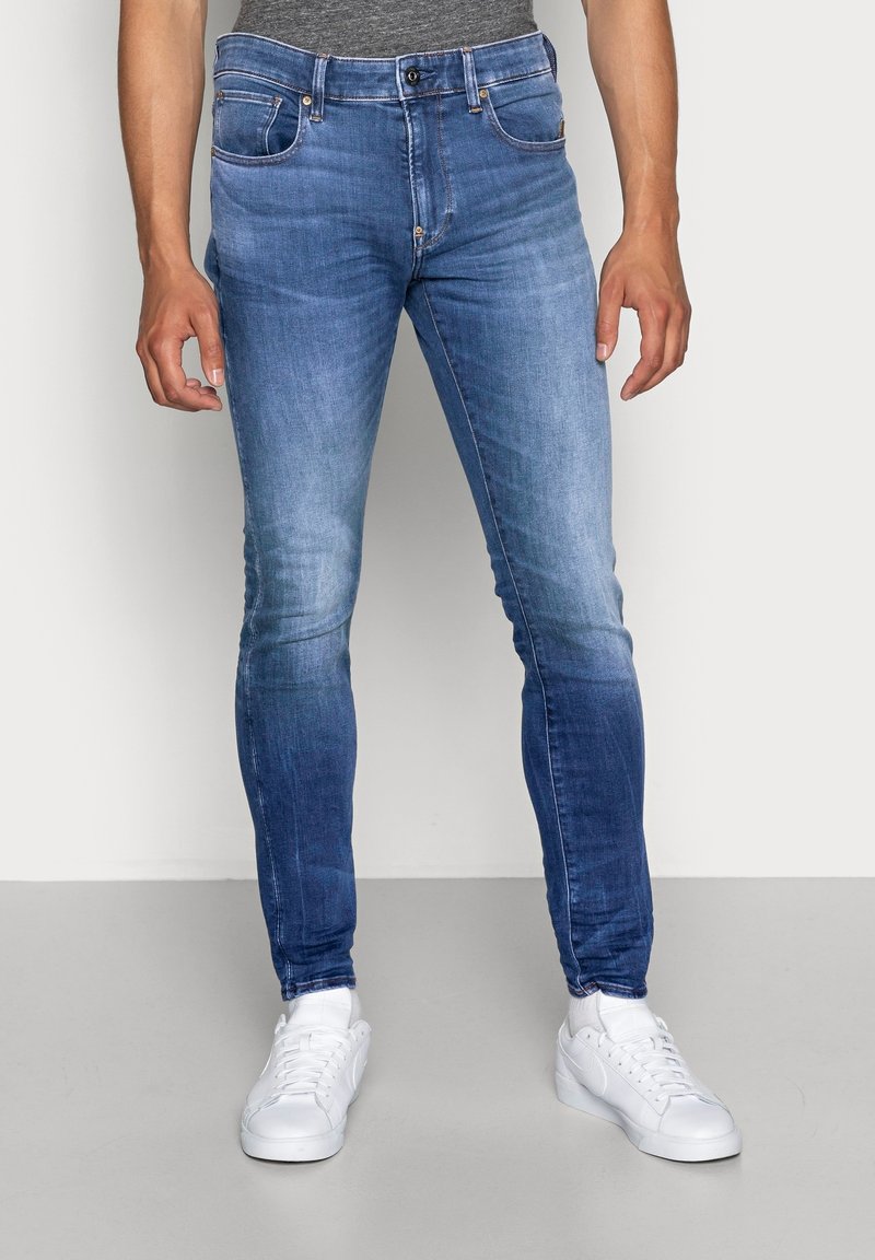 Homme portant un jean bleu délavé slim fit et des baskets blanches, debout devant un fond clair uni.