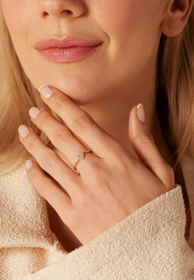 Gouden ring met een hartvormig ontwerp, met een cluster van kleine steentjes. Vingers hebben een lichte roze nagellak, tegen een gestructureerde, beige stof.
