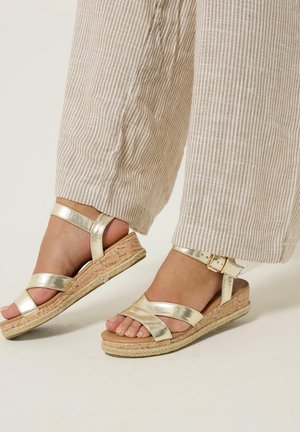Voeten met metallic gouden sleehak sandalen met enkelbandjes, kurken zolen en beige broek met wijde pijpen en fijne strepen.