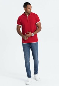 Polo rosso con dettagli bianchi, maniche corte, colletto con due bottoni, abbinato a jeans blu e scarpe da ginnastica bianche, in piedi davanti a uno sfondo neutro.