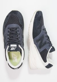 Tenisky New Balance mají tmavě námořnický svršek s panely z semiše a síťoviny, kontrastní šedé detaily a lehkou bílou mezipodešev.