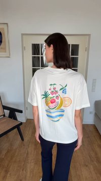 Femme debout à l'intérieur, cheveux foncés, portant un t-shirt blanc avec des illustrations colorées de fruits dans le dos, un pantalon bleu marine et des chaussures blanches.