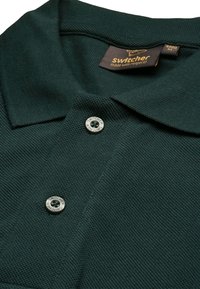 Polo shirt verde scuro con tessuto testurizzato, patta con tre bottoni caratterizzata da bottoni in metallo, e etichetta "Switcher" sul colletto.