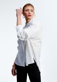 Camicia bianca a maniche lunghe con colletto classico, caratterizzata da bottoni decorativi e una vestibilità ampia. Indossata con pantaloni scuri e orecchini a cerchio.