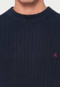 Pulover tricotat navy cu textura în caneluri, cu guler rotund, având un mic accent logo roșu. Textura pare moale, iar designul este ajustat.