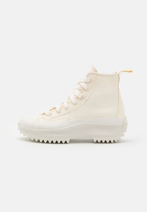 Zapatilla beige de lona de caña alta con suela blanca gruesa y acanalada, cordones blancos y una lengüeta trasera, vista de lado.
