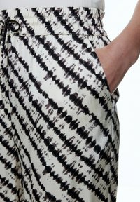 Leichte Hosen aus einem cremefarbenen Stoff mit dunklem Batikmuster. Verfügt über einen elastischen Bund und Seitentaschen. Weiche Textur.