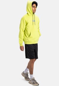 Yellow hooded sweatshirt with a front pocket, paired with black shorts. Model wears grey sneakers and white socks with a purple pattern.