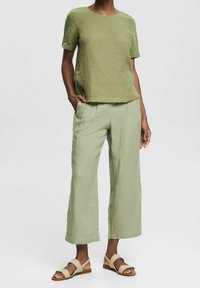 Mujer usando una blusa de lino de manga corta de color verde claro, pantalones cropped de pierna ancha a juego y sandalias de dedo abiertas color beige, de pie con una mano en el bolsillo.