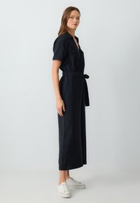 Combinaison noire à manches courtes avec col, taille cintrée et jambes larges. Stylée avec des baskets blanches sur un fond clair.