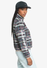 Fleece jacket with a multicolored geometric pattern in purple, blue, and green, featuring a high collar and white logo on the sleeve.