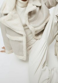 Chaqueta de imitación de piel de cream con forro de pelo, cuello con solapa y bolsillos, combinada con un cuello alto de color crema claro y pantalones lisos beige.