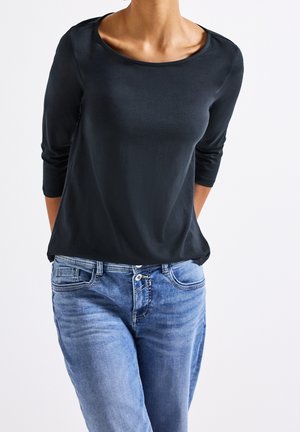 Personne portant une chemise noire à manches longues et un jean en denim bleu, debout devant un fond blanc uni.