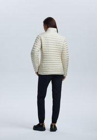 Lys beige puffervest med horisontale syninger, høj krave og glat tekstur, parret med sorte tapered bukser og sorte sneakers.