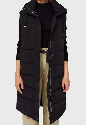 Woman wearing a long black padded sleeveless vest over a black sweater and beige pants with a black belt, standing against a plain background.
