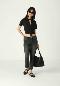 Mulher vestindo um top cropped preto franzido, jeans cinza escuro, sapatos pretos, óculos de sol e segurando uma grande mala preta, de pé contra um fundo branco.