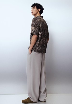 Nuori mies, jolla on kiharat hiukset, pukeutunut kuvioituun ruskeaan lyhythihaiseen paitaan, leveisiin vaaleanharmaisiin housuihin ja ruskeisiin slip-on-kenkiin, seisoo sivuttain.