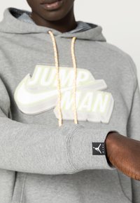 Sweat à capuche gris en mélange de coton. Présente un grand graphique "JUMPMAN" en blanc et jaune. Comprend des cordons multicolores et un patch logo.