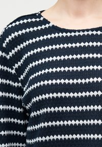 Gros plan sur un pull en maille rayé en zigzag bleu marine et blanc porté par une personne, montrant la partie supérieure de la poitrine et de l'épaule.