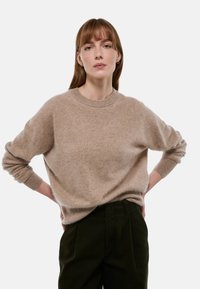 Pull beige oversize avec un col rond et des manches longues, fait d'un matériau doux et duveteux, associé à un pantalon vert foncé.