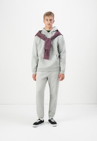 Ensemble de survêtement gris avec un sweat à capuche et un jogging, surmonté d'une chemise maroon à carreaux nouée autour du cou. Baskets noires avec des semelles blanches.