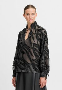 Blusa nera trasparente con frange nere testurizzate a motivo foglia, colletto alto e dettagli al collo con nodo. Maniche lunghe con legature sui polsini.