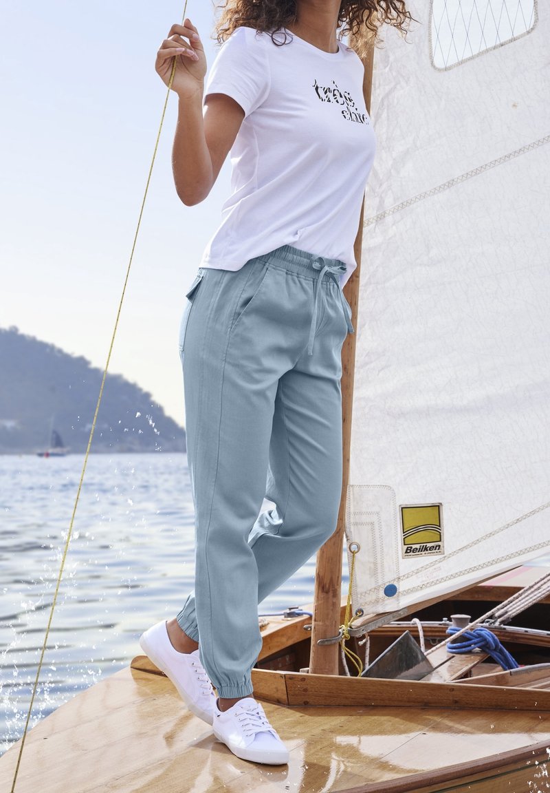 Nainen seisoo puuvenesataman kansilla pitäen köyttä, päällään valkoiset lenkkarit, vaaleansiniset housut ja valkoinen t-paita, jossa on teksti.