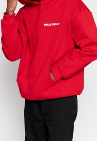 Persona che indossa un felpa rossa con la scritta "MULTIPLY" sul petto, una mano nella tasca della felpa, abbinata a pantaloni neri su uno sfondo semplice.