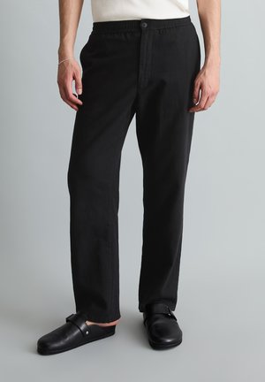 Homme portant un pantalon noir droit avec une taille élastique et des chaussures noires à enfiler sur fond uni.