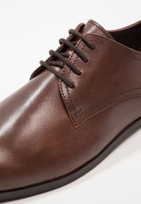 Bruna läderdojor med en slät yta, som har sex öglor, mörka snören och subtila sömmar längs sidorna.