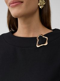 Vrouw in een zwarte top met een abstracte gouden broche op de kraag en bijpassende gouden sculptuur oorbellen, met een zichtbare hals en onderzijde van het gezicht.