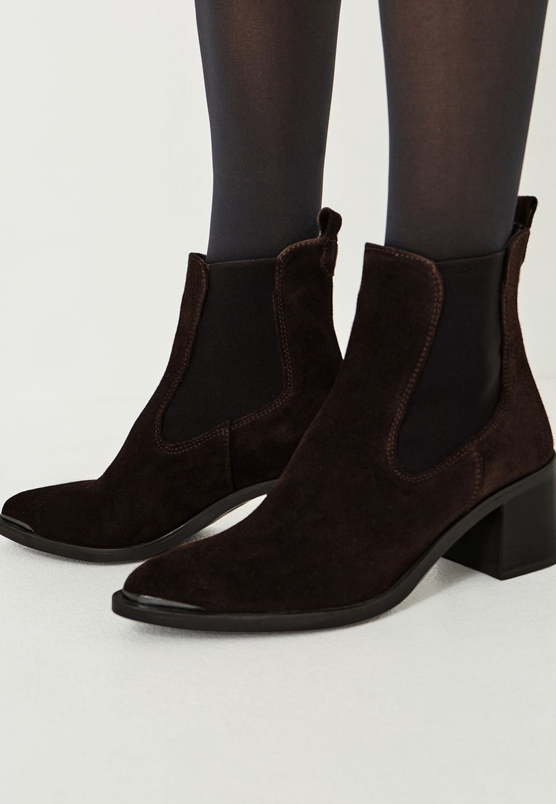 Bottines en suède marron foncé à talons blocs, portées avec des collants noirs sur fond clair.