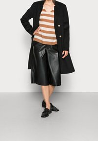 Schwarzer langer Mantel, cremefarben- und braun gestreifter Strickpullover, schwarze Leder-Culottes und schwarze Loafers, die Texturen und geschichtete Stile zeigen.