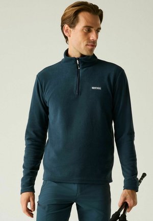 Man wearing dark blue fleece pullover holding folded trekking poles, looking sideways against plain background.