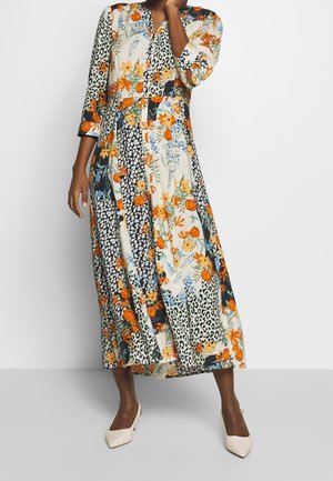 Femme portant une longue robe patchwork à imprimé floral et léopard, manches trois-quarts et chaussures blanches à bout pointu.