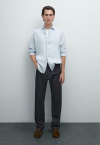 Camicia azzurra in lino con bottoni, jeans in denim scuro, scarpe in suede marroni. Modello in piedi con le mani in tasca su uno sfondo neutro.