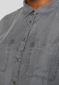 Camicia in chambray grigia con colletto a pulsante, patta frontale e due tasche sul petto. Presenta un tessuto morbido e testurizzato e bottoni metallici.