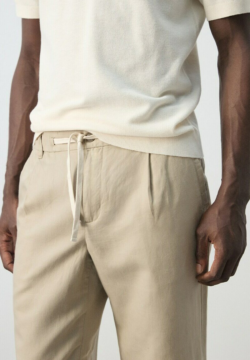 Man draagt beige trekkoordenbroek en een licht beige textuur korte mouwen overhemd, staand met ontspannen handen langs de zij.