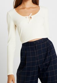 Kerman ribbattu pitkähihaine paita, jossa on pyöreä kaula-aukko ja nauhakiinnitys edessä, yhdistettynä tummansinisiin ruudullisiin korkeavyötäröisiin housuihin.