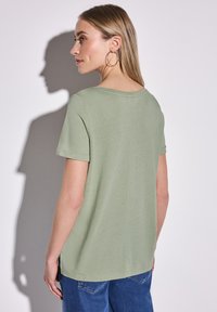 Chemise verte claire à manches courtes en tissu doux. Présente un décolleté en forme de scoop et des fentes sur les côtés au niveau de l'ourlet. Portée avec un jean bleu.