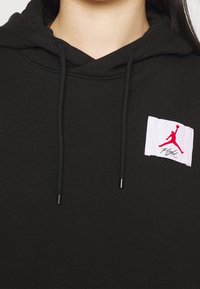 Sudadera negra de tejido suave, con un parche blanco que cuenta con un logotipo rojo de Jordan y texto, cordones con puntas metálicas y un ajuste relajado.