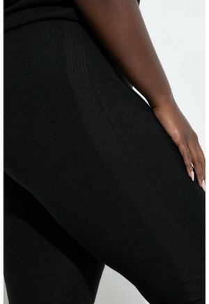 Mørkhudet person iført sorte tætsiddende bukser, hånd hviler på hoften, hvilket fremhæver stoftekstur og syningsdetaljer langs låret.