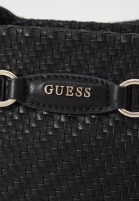 Czarna pleciona torebka z teksturowego materiału, skórzanymi akcentami i wyraźnym srebrnym logo "GUESS" na czarnym pasku.