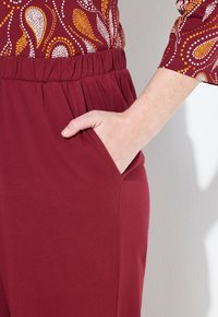 Bordeauxkleurige broek met zijzakken, voorzien van een elastische taille en zachte textuur, gecombineerd met een gemusterde top in gele en witte tinten.