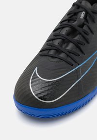 Zwart voetbalschoen met een gestructureerde bovenkant, met een opvallende zilveren Nike-swoosh en een blauwe rubbersole. Veters en veteroogjes zijn inbegrepen.