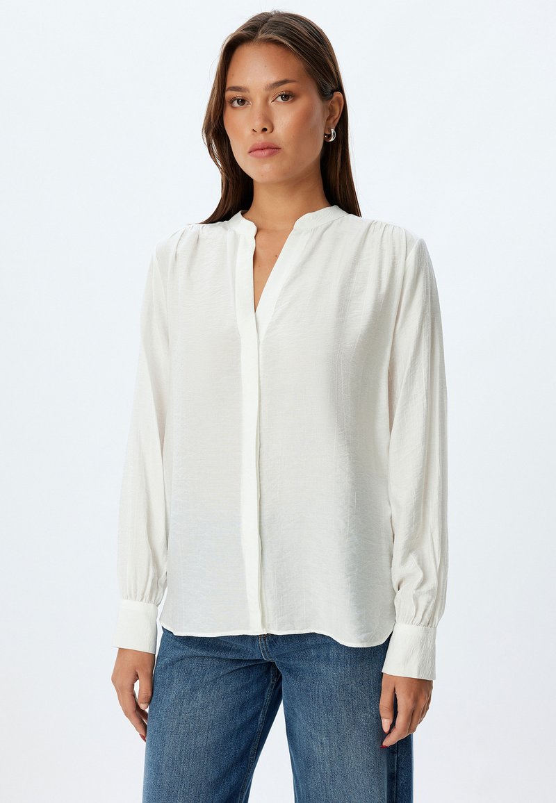 Witte blouse met lange mouwen, een v-hals en knoopsluiting, gemaakt van zachte, gestructureerde stof en licht gepofte schouders. Gekleed met blauwe jeans.