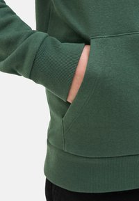 Sweatshirt en coton vert avec des poignets côtelés, doté d'une poche frontale et d'une texture douce. Vue rapprochée d'une main dans la poche.