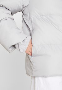 Světle šedá pé�řová bunda s hladkým, lesklým povrchem, která má skrytou kapsu a měkký šedý pletený manžet, viditelný pod bundou.