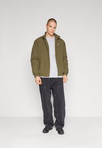 Chaqueta bomber verde oliva con puños elásticos, camiseta gris debajo, pantalones vaqueros negros holgados con bolsillos frontales y zapatos negros. Fondo neutro.