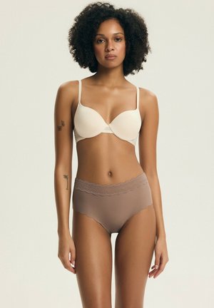 Kobieta o kręconych włosach stoi ubrana w beżowy biustonosz i majtki z fioletowym koronkowym wykończeniem, z tatuażami motyla i palmy na lewej ręce.